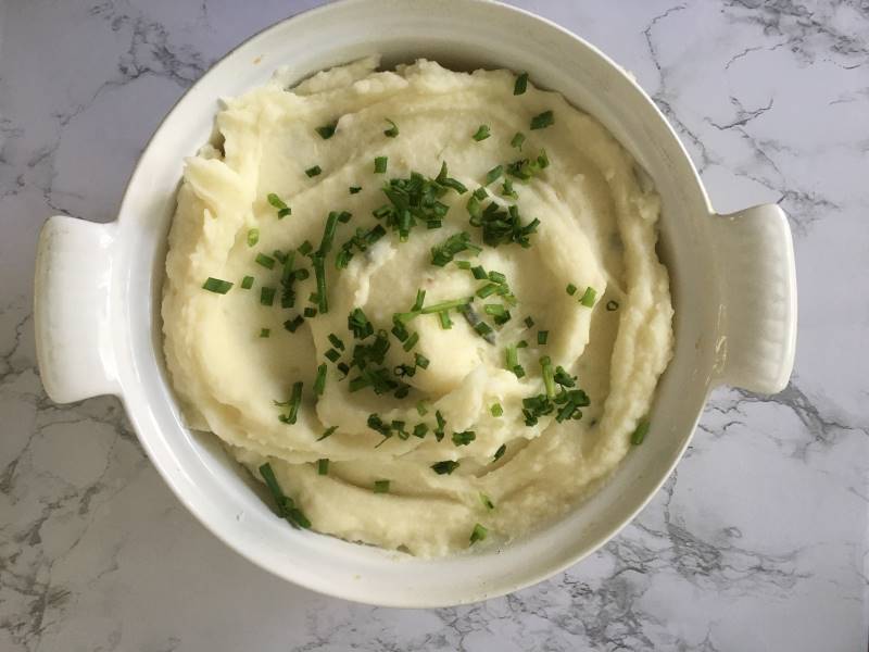 Finished bowl of garlic and herb mashed potatoes, sprinkled with chives, on a quartz cutting board