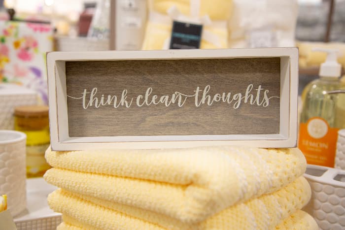 Rustic farmhouse bathroom sign with "think clean  thoughts" in white lettering, placed on stack of pale yellow towels with out of focus bathroom in the background.