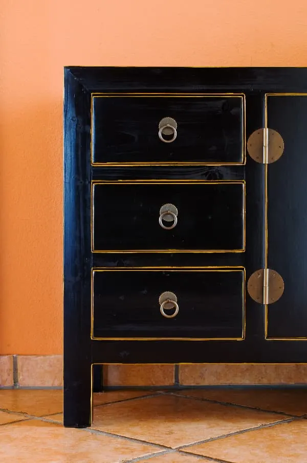 A partial view of a dark wooden dresser with thin gold lines painted around the edge of the front of the drawer faces and door along the openings for the drawers and door.