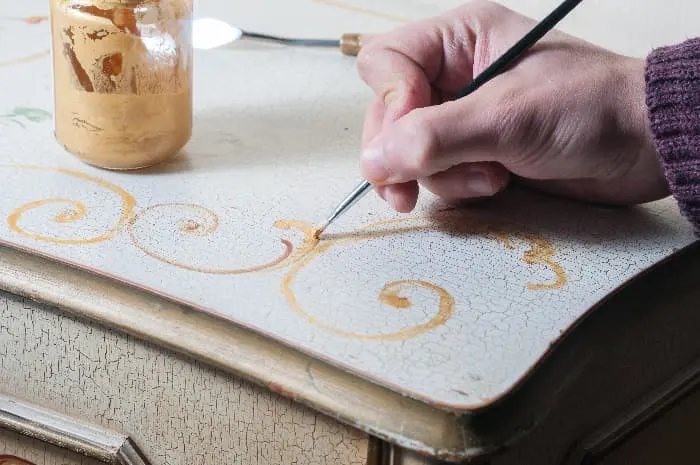 A closeup of a hand painting a gold curlicue design in the corner on the top of a white painted dresser.