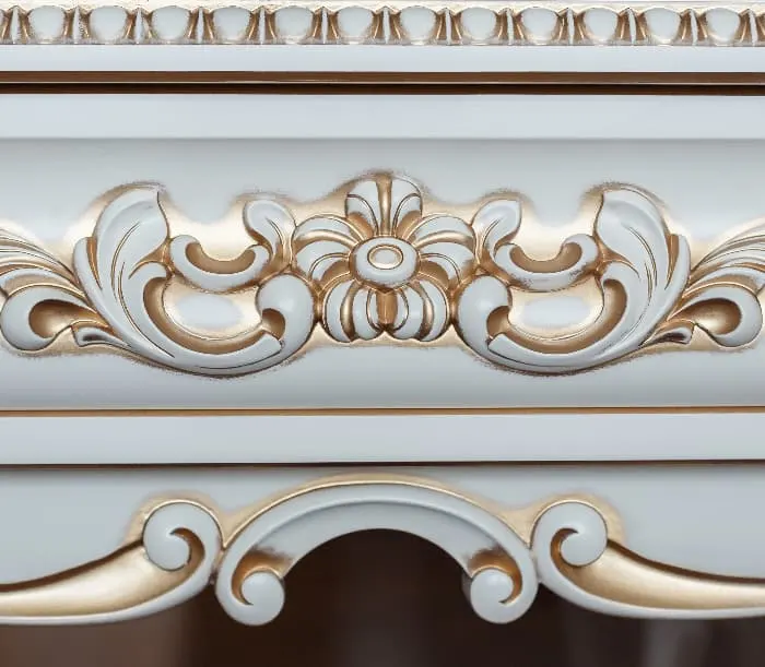 Closeup of gold paint-highlighted ornate detailing on a white painted dresser.