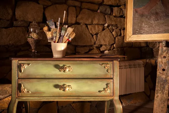 An antique painted dresser with an olive dressed paint job and wooden designed added to the front of the drawers and around the knobs, a stoneware vase holding paintbrushes and a glass jar on top of the dresser, against a stone wall.