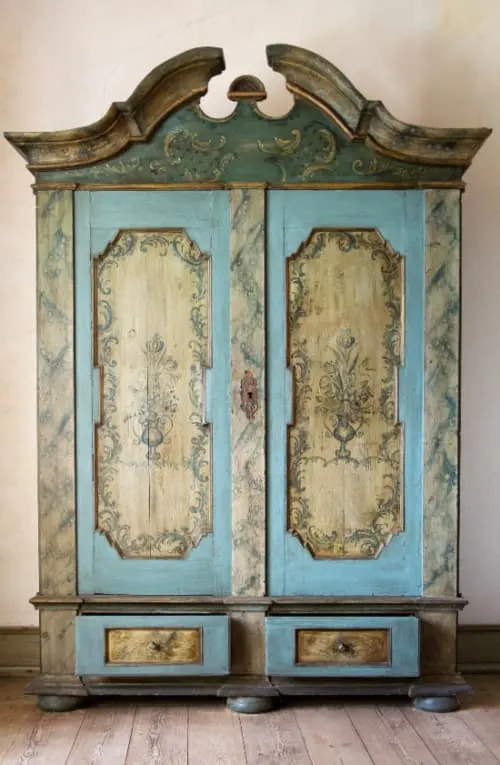 A ornate armoire with blue paint and subtle blue floral designs painted on inlaid panels on the doors and the drawers.