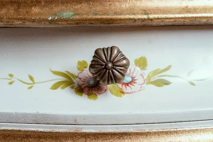A closeup of stenciled floral elements behind a decorative floral motif knob on a white painted dresser drawer.