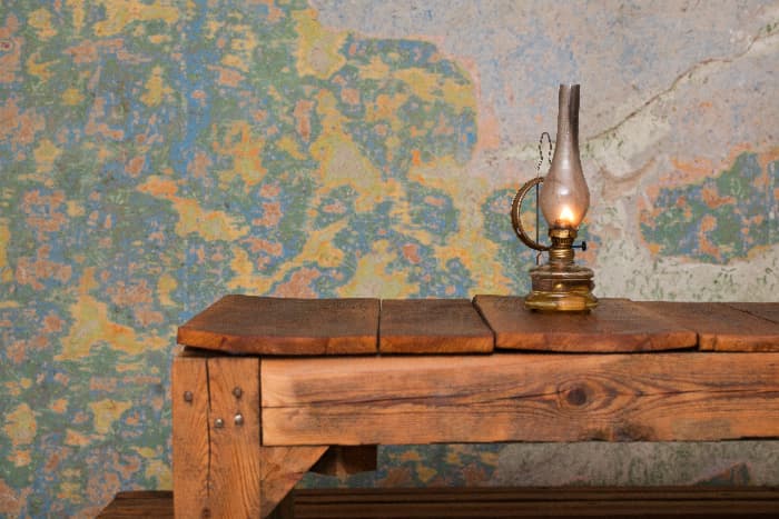 Medium-stain table made of warped planks against wall with peeling wallpaper.