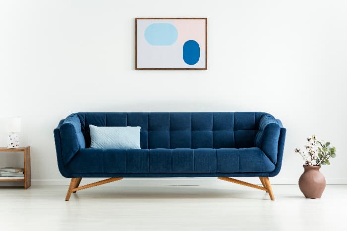 A modern style sofa in navy blue fabric in a white living room with a piece of blue abstract art hung above the sofa -- an example of understate and elegant blue furniture.