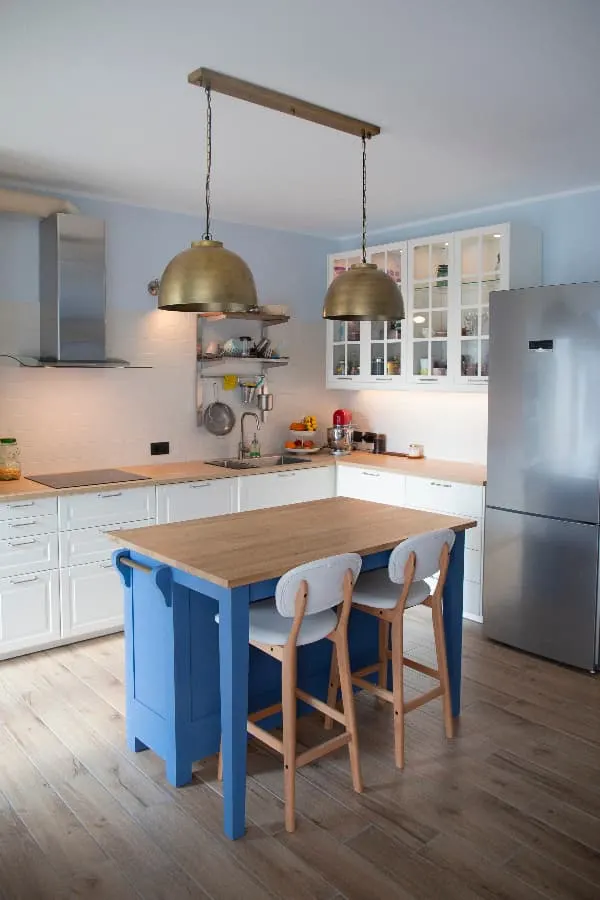 A white kitchen with butcher block counters, glass front cabinets, and stainless appliances with a kitchen island painted a primary blue color -- a blue island brings blue furniture into the kitchen.