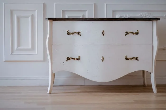 White painted dresser with black top, brass hardware and curved lines against a white wall and light wood flooring.