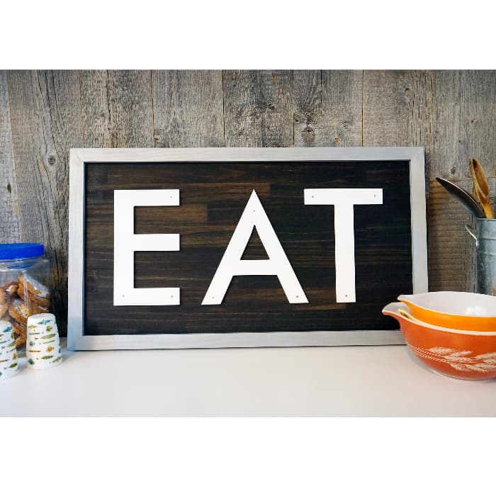 A metal-framed farmhouse kitchen sign with the word "EAT" in bold white letters on a dark wooden background.  The sign is on a white counter top propped against a rustic wooden plank wall.