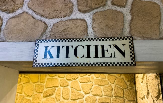 A farmhouse kitchen sign with black and white checkerboard along the edges with "KITCHEN" in black letters. The sign is hung on a wooden beam in a stone wall.