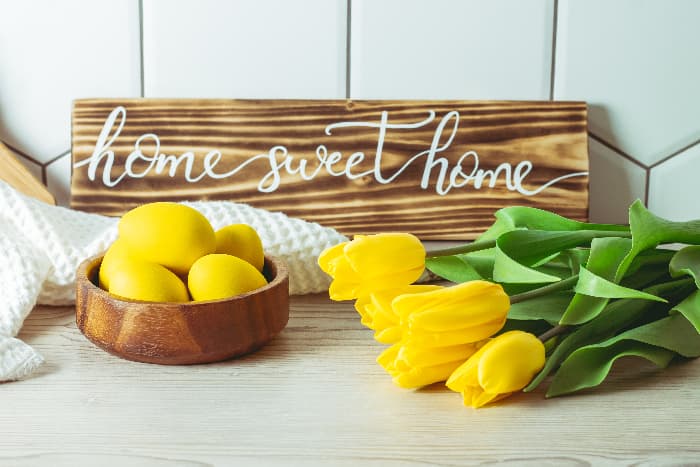 A wooden plank farmhouse kitchen sign with "home sweet home" in white script lettering.  The sign is propped on a white-washed wooden counter against a white tiled wall, with yellow tulips and a wooden bowl of yellow-colored eggs in front of the sign.