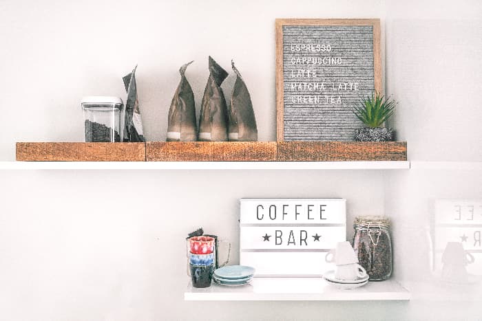 A white farmhouse sign with "COFFEE BAR" in black letters.  The sign is on a white floating shelf with beneath a longer white floating shelf with various coffee-related items on them.