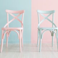 Two painted chairs, one painted pale pink against a pale blue background, the other painted pale blue against a pale pink background