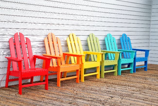 A multi-color grouping of painted chairs -- six Adirondack chairs against a clapboard siding wall with each one painted a different color (red, orange, yellow, pale green, aqua, and blue).