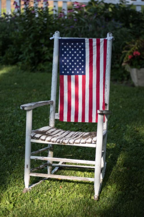 An antique rocking chair with white distressed paint and an American flag on the back of the chair -- a patriotic painted chair theme.