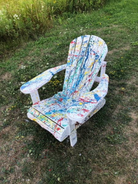 A white painted Adirondack chair with splattered paint in primary colors.