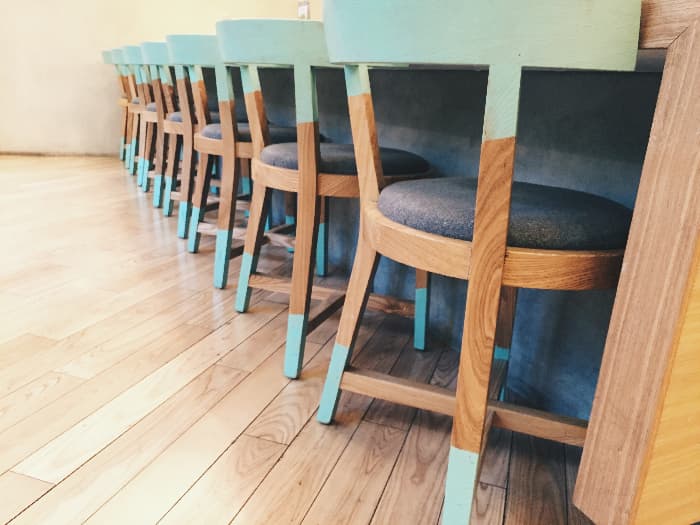A row of painted chairs at a long bar/counter that have blue paint on the backs of the chairs and also at the bottoms of the legs -- all other parts of the chair are stained wood.