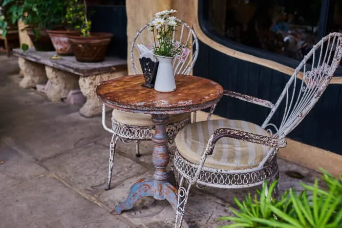 Outdoor patio/porch scene with a round painted table that has been distressed and given a patina to make it appear aged. Two distressed painted metal chairs with striped cushions are on either side of table.
