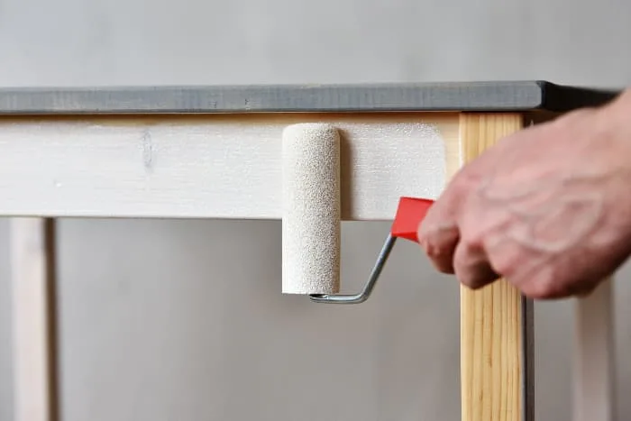 Closeup of a man's hand painting the base of a table white with a small paint roller.