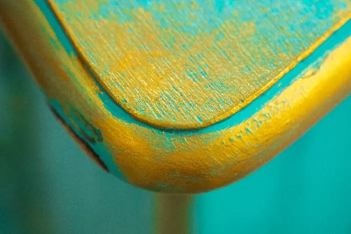 Closeup of a painted table top that has turquoise paint showing through a bright yellow coat of paint.