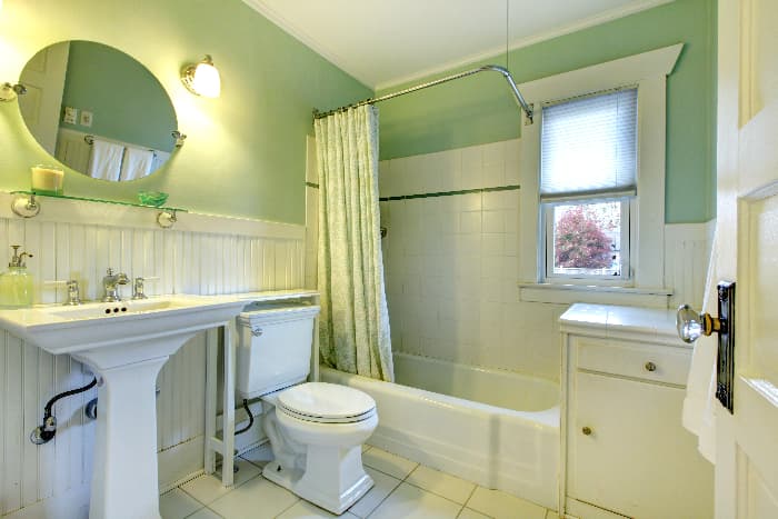 Bathroom with pale green wall color. shower curtain with a white and pale green design, white wainscotting, and woodwork white shower and floor tile, white bathroom fixtures, and a white cabinet.