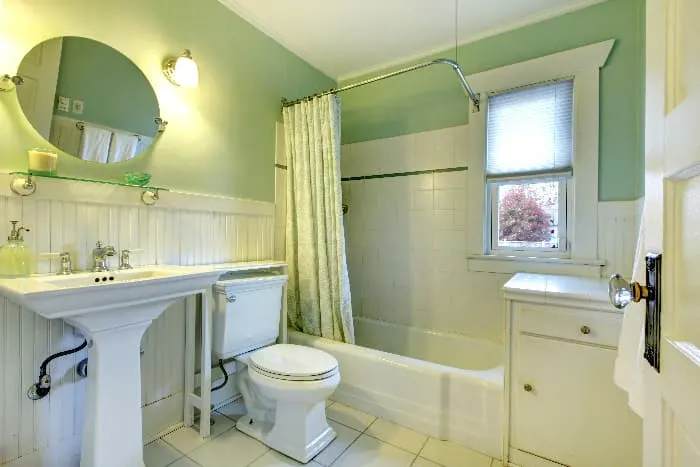 Bathroom with pale green wall color. shower curtain with a white and pale green design, white wainscotting, and woodwork white shower and floor tile, white bathroom fixtures, and a white cabinet.