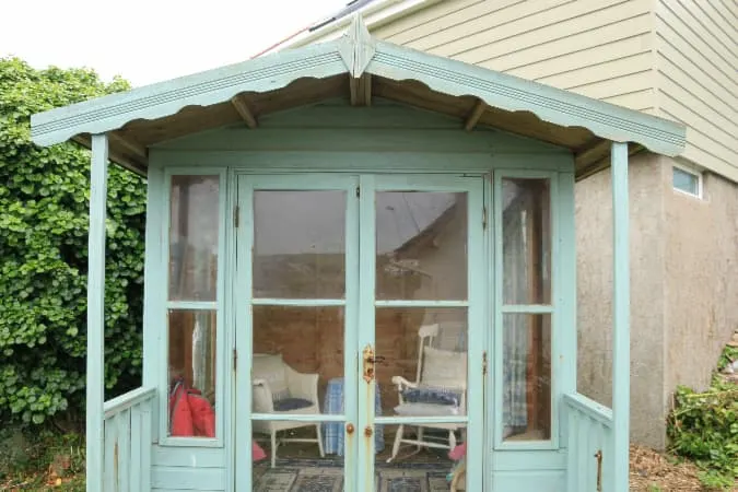 Decorative shed in pale green color with small covered front porch, French doors, and white furniture viewed through door window panes.