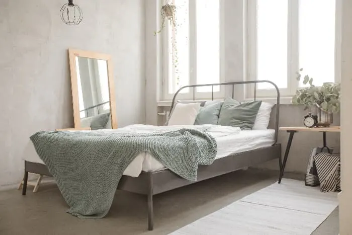 A pale green throw blanket n a modern bed with a gray metal frame located beneath tall, uncovered windows in a white bedroom.