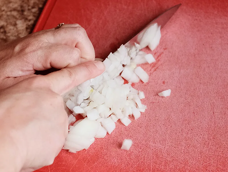 Hands using a knife to dice onions on a red cutting board.