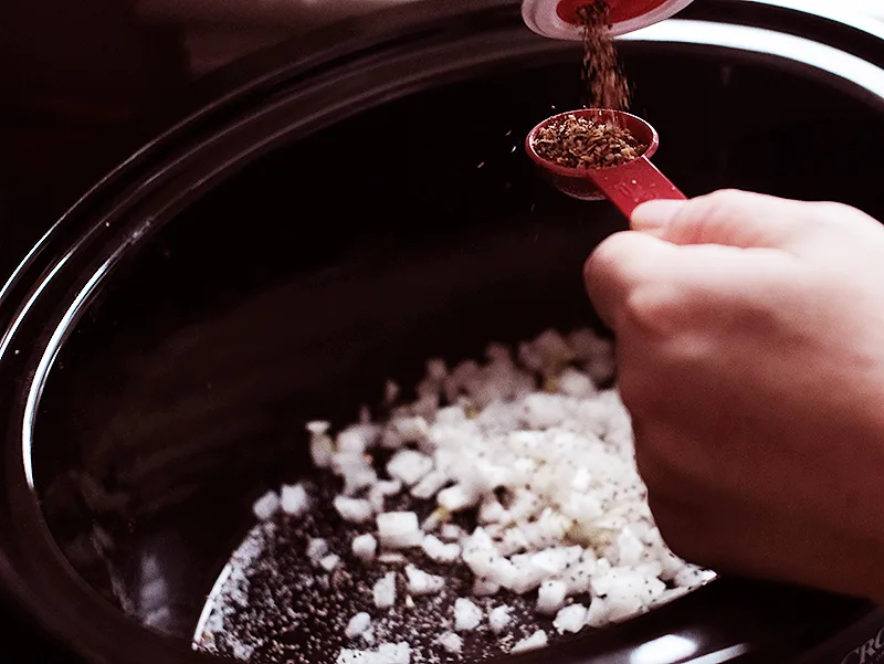 Hands pouring steakhouse seasoning mix into a measuring spoon over diced onions in a slow cooker.