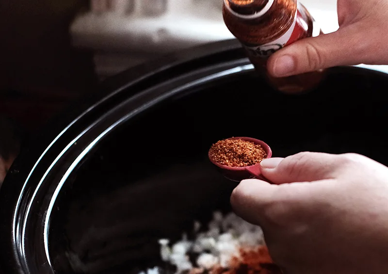 Hands measuring out Tajin spice mix over a slow cooker.