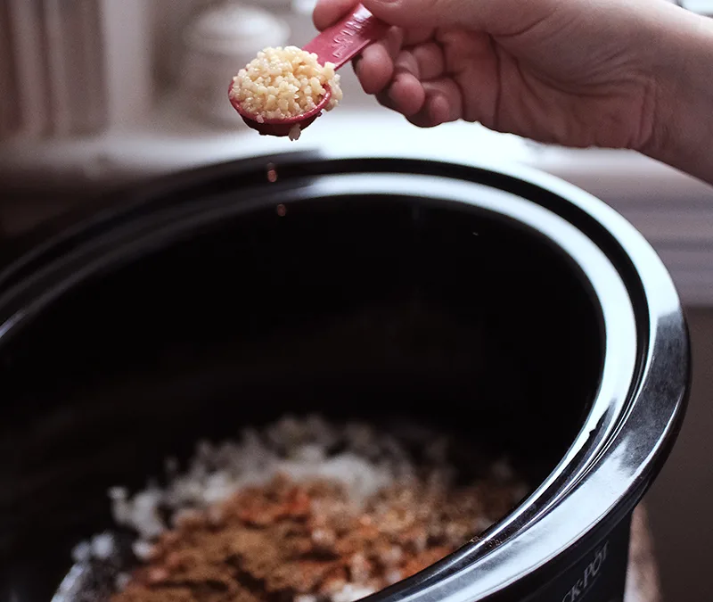 Hands adding a heaping measuring spoon of minced garlic to ingredients in a slow cooker.