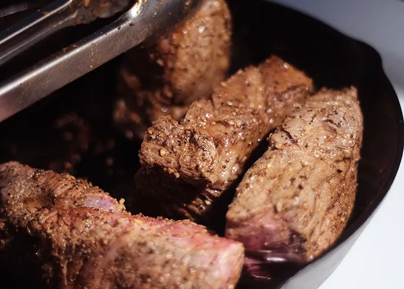 Tongs turning pieces of chuck roast browning in a skillet.