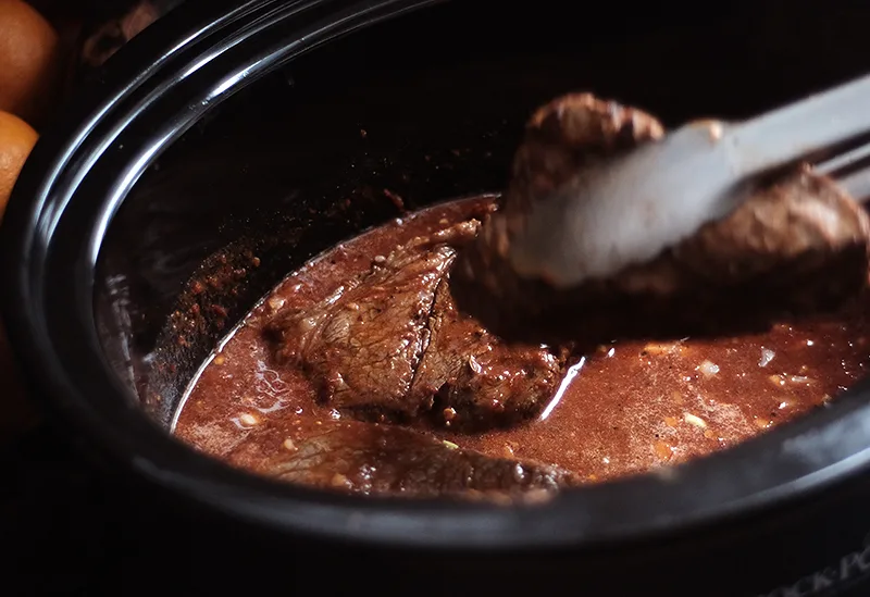 Tongs placing browned chuck roast pieces in slow cooker.