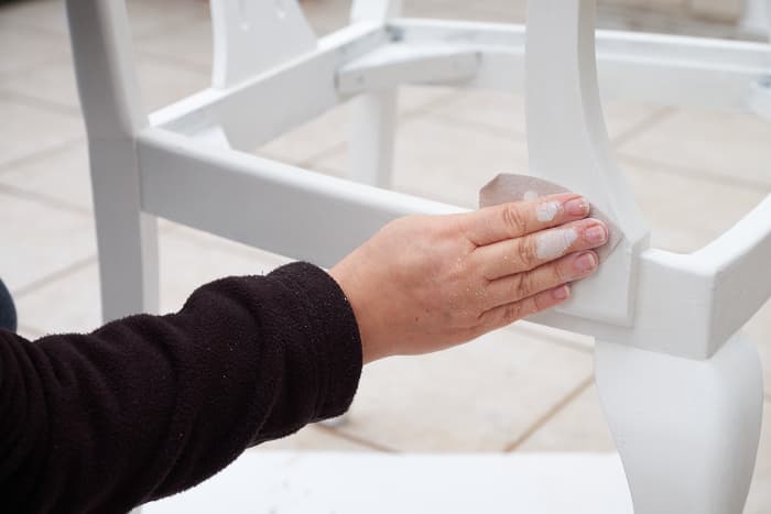 Closeup of woman's hand sanding white painted furniture.  Sanding between coats will help give you a smooth finish with chalk paint.