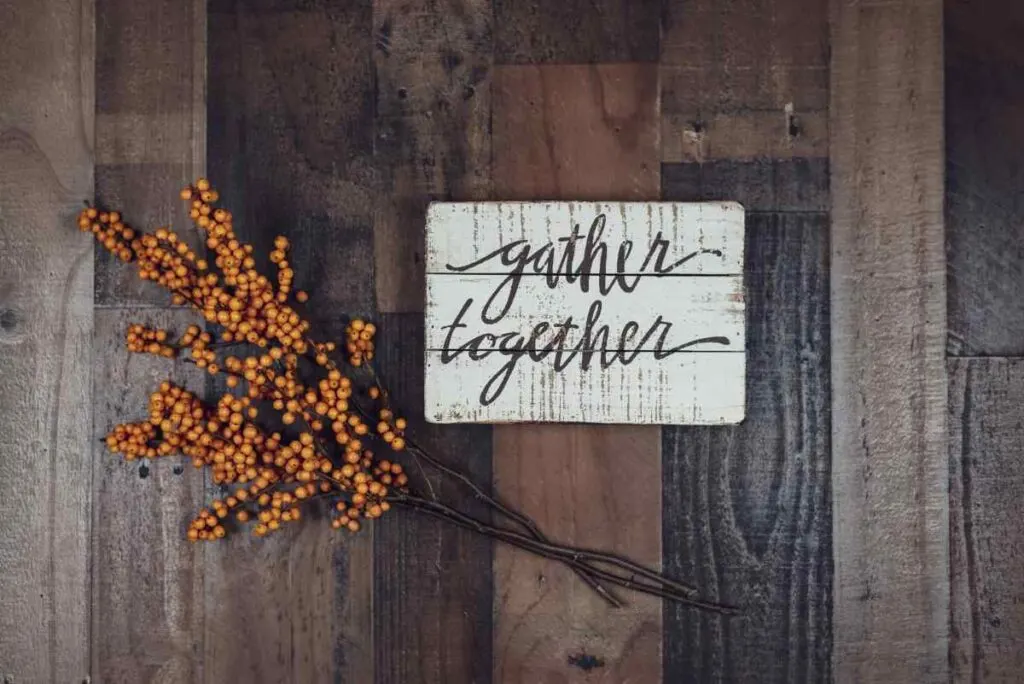 The words Gather Together painted on a white distressed wood sign with berries