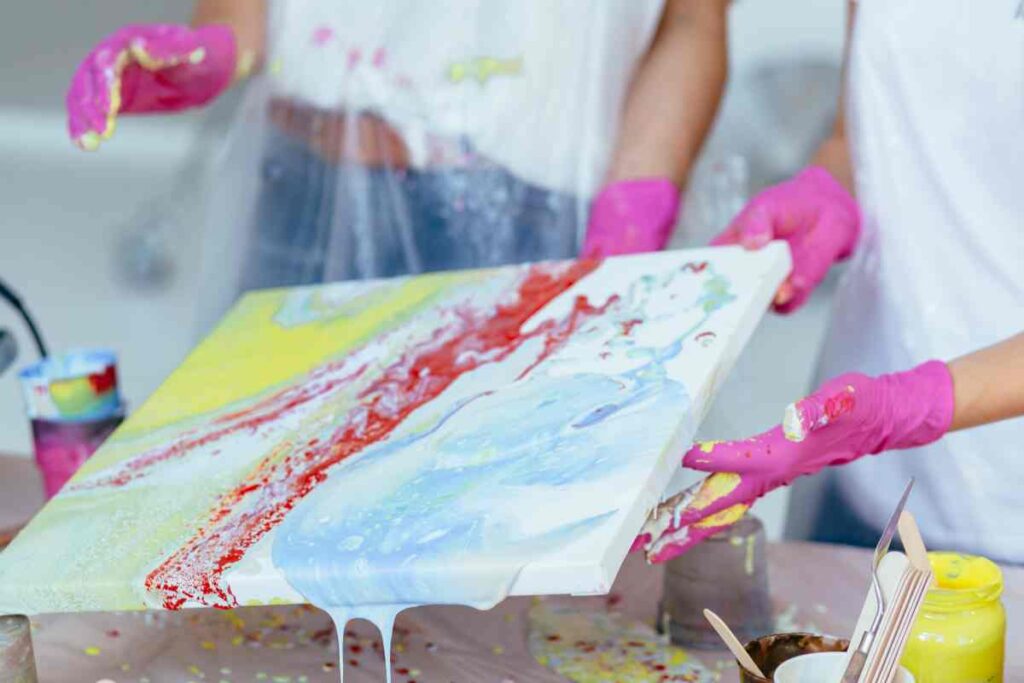 Hands holding a colorful canvas with paint pouring supplies on the table