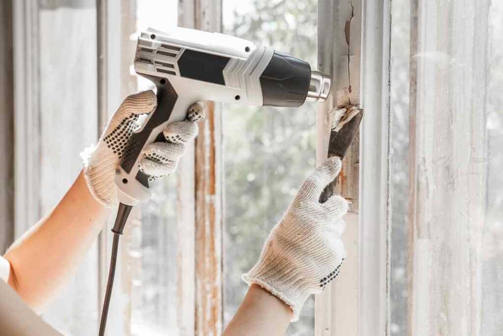 Using a heat gun and scraper to remove white paint from a window frame
