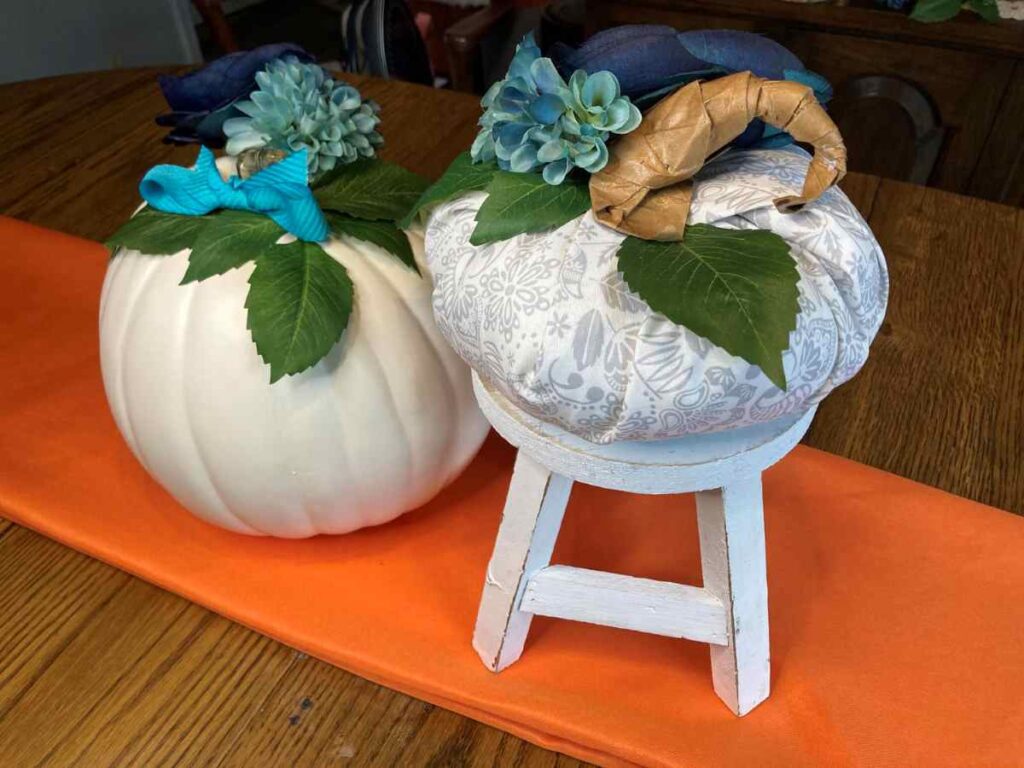 Grey and white fabric pumpkin and white faux pumpkin decorated with flowers displayed on an orange runner