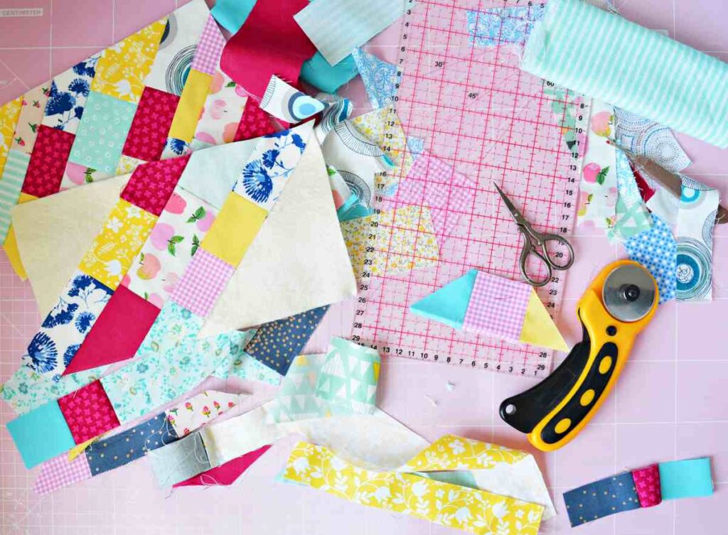 Quilting supplies including a rotary cutter, scissors, quilting ruler, scrap fabric, and completed quilt block