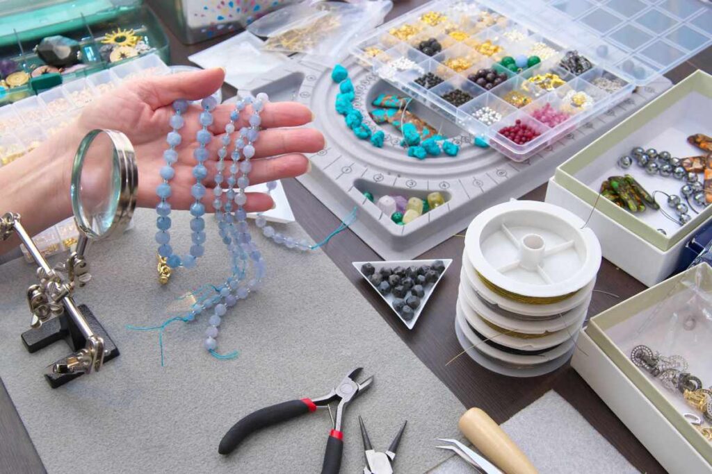 A woman holding three beaded necklaces with a table covered in jewelry making supplies in the background