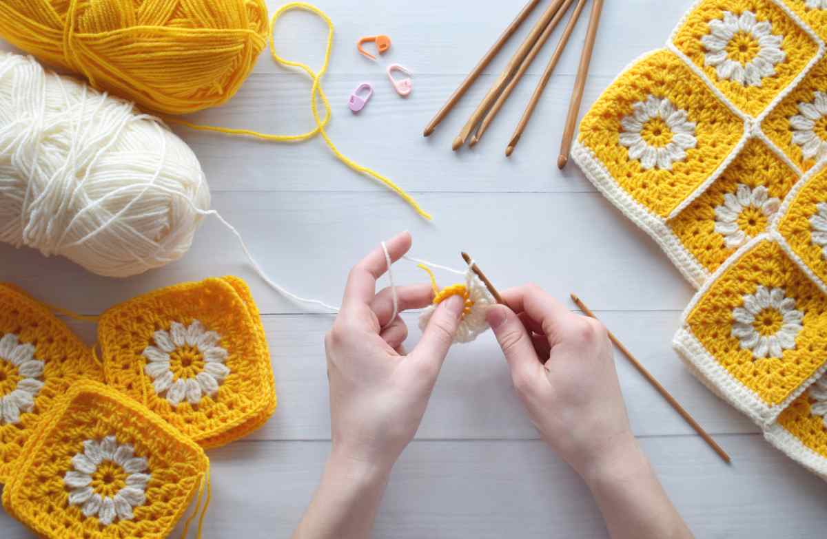 Woman hands with crochet hooks, yellow and white yarns, and flower crochet patterns